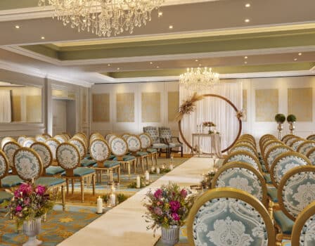 Elegant wedding ceremony steeing with ornate chandeliers, gold chairs, and floral arrangements in luxurious hotel ballroom.