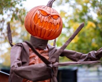 Halloween-themed scarecrow with carved pumpkin head amidst autumn trees at outdoor hotel garden.