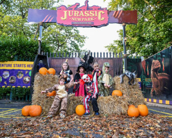 Guests in Halloween costumes around hay bales at Jurassic-themed park entrance with pumpkins and vibrant signage.
