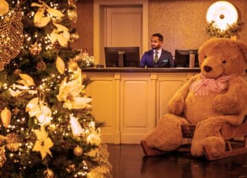 Hotel lobby with festive Christmas tree, plush teddy bear, and friendly receptionist, welcoming guests in a cosy atmosphere.