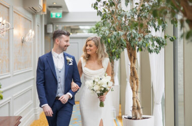 Bride and groom walk arm in arm through an elegant indoor corridor at Newpark Hotel Kilkenny, smiling at each other as the bride holds a white bouquet surrounded by greenery and soft natural light.