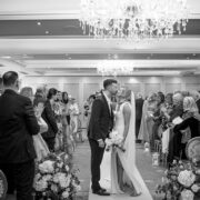 Bride and groom share their first kiss at the end of their wedding ceremony in a chandelier-lit ballroom at Newpark Hotel Kilkenny, surrounded by applauding guests seated along the aisle.