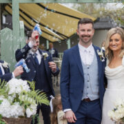 Bride and groom smile together outside Newpark Hotel Kilkenny as wedding guests blow bubbles in celebration, with the couple holding a bouquet and standing beneath a covered outdoor walkway.