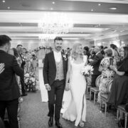 Newly married bride and groom walk hand in hand back down the aisle after their wedding ceremony at Newpark Hotel Kilkenny, smiling as guests applaud in a chandelier-lit ballroom.