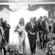 Bride walks down the aisle with her father during her wedding ceremony at Newpark Hotel Kilkenny, as guests stand and watch in an elegant chandelier-lit ballroom.