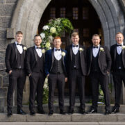 Groomsmen in suits pose by hotel's elegant stone arch, adorned with lush floral decorations, ideal for weddings.