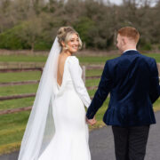 Bride and groom walking hand-in-hand on scenic path with lush greenery and rustic wooden fence at countryside hotel venue.