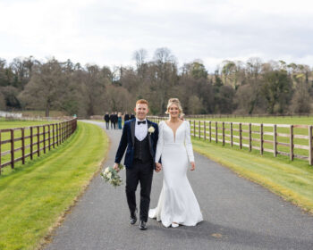 Elegant couple walking on scenic countryside path, surrounded by lush greenery and rustic fencing at luxury hotel venue.