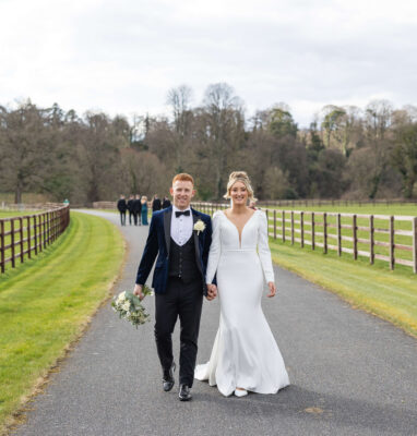 Elegant couple walking on scenic countryside path, surrounded by lush greenery and rustic fencing at luxury hotel venue.