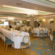 Elegant hotel ballroom with chandeliers, ornate tables set for an event, floral arrangements, and plush seating.