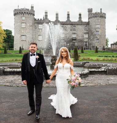 Elegant wedding couple in front of historic castle with fountain, lush gardens, perfect for memorable hotel celebrations.