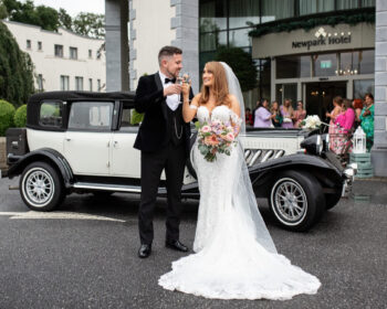 Newlyweds celebrating with champagne in front of a vintage car at Newpark Hotel entrance, perfect for weddings.