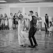 Elegant ballroom wedding dance in spacious hotel venue with wooden flooring, surrounded by smiling guests.