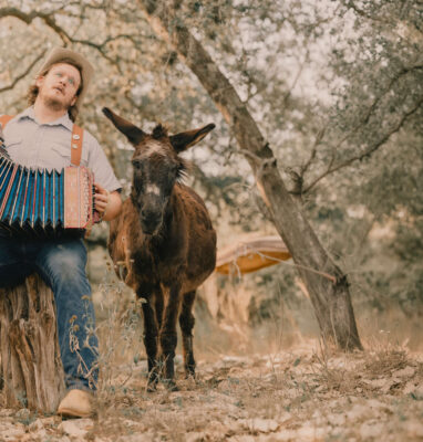 Guest enjoys playing accordion next to a friendly donkey under shaded oak trees, showcasing rustic charm and outdoor leisure.
