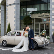 Newlyweds share a kiss outside Newpark Hotel, surrounded by elegant architecture and a classic car, perfect for weddings.