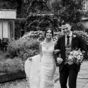 Happy couple in wedding attire strolling through lush hotel courtyard with flowers and ivy, offering unique wedding venue experience.