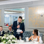 Elegant wedding banquet room with smiling guests and a groom speaking, set in a luxurious hotel setting.