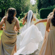 Bride in white gown joyfully walks with bridesmaids in lush garden path at scenic hotel venue.