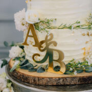 Elegant white wedding cake with floral accents and gold initials on rustic wooden stand at luxury UK hotel venue.