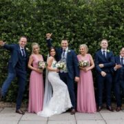 Happy wedding party poses in elegant attire against lush greenery at hotel garden.
