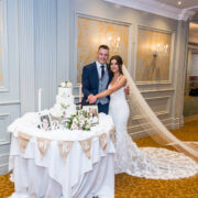 Elegant wedding reception with smiling couple cutting cake in luxurious hotel ballroom adorned with soft gold accents.