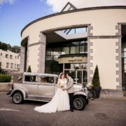 Elegant wedding couple with vintage car in front of stylish hotel entrance with modern architecture and colourful mural.