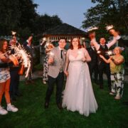 Newlyweds celebrate with sparklers surrounded by friends in lush garden beside elegant hotel gazebo at twilight.
