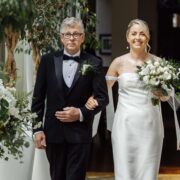Bride being walked down to her elegant wedding ceremony with floral décor in luxury hotel setting, ideal for romantic occasions.