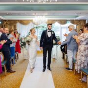 Elegant wedding ceremony in a spacious hotel ballroom with chandelier, lush decor, and guests applauding the couple.