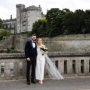 A joyful couple in wedding attire smiles on a historic stone bridge with an enchanting castle and lush greenery backdrop.