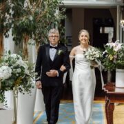 Smiling bride in elegant dress walks with a partner through lush, floral hotel hallway.