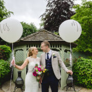 Couple celebrates wedding in lush garden gazebo with balloons, surrounded by vibrant greenery and elegant decor.