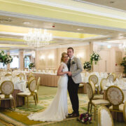 Elegant ballroom with chandeliers, round tables, and floral centrepieces, ideal for weddings. Couple stands embracing joyfully.