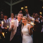 Newlywed couple celebrates with sparklers at an elegant wedding venue. Smiling guests in smart attire join the festivities.