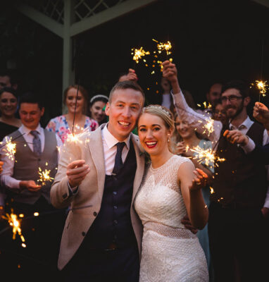 Newlywed couple celebrates with sparklers at an elegant wedding venue. Smiling guests in smart attire join the festivities.