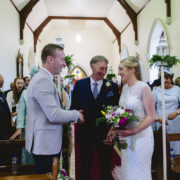 Bride and groom exchanging vows in elegant church with guests smiling in approval, vibrant floral arrangements visible.