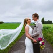 Bride and groom smiling on a lush countryside path, her veil flowing in the breeze, surrounded by vibrant green fields.