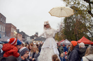 Entertaining street performer on stilts at lively festival with colourful stalls and crowd near cosy hotel.